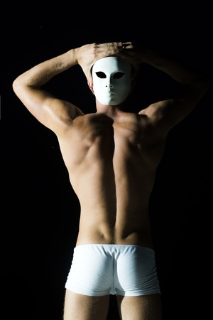 Back view of handsome sexual young man with straight body and beautiful bottom in white underwear holding white mask near head standing in studio on black background, vertical pictureの写真素材