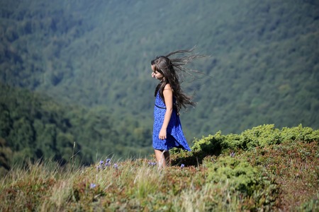 Little brunette child girl in blue lace summer dress looking away standing in mountain valley with deep dry grass sunny windy day outdoor on natural background, horizontal pictureの写真素材