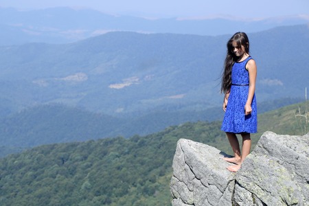 One brunette pensive girl with long hair in blue lace dress standing on stone rock cliff looking down on beautiful landscape sunny day outdoor natural blue sky background copyspace, horizontal photoの写真素材