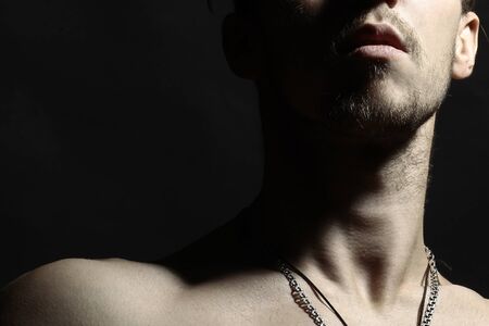 Portrait of handsome wellbuilt young naked white brunette with mustache beard and silver chain on neck in shadow on black background, horizontal photoの写真素材
