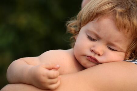 Closeup of one cute relaxing lovely small male child with blonde curly hair sleeping on human hands of parent in sun light outdoor on natural green background, horizontal pictureの写真素材