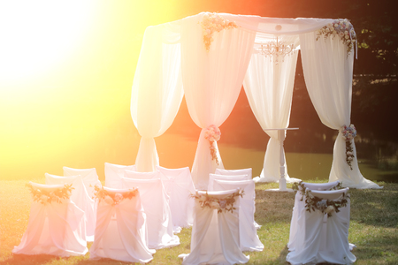 Beautiful decorated with light white chiffon chandelier and bouquets of roses pastel colors wedding pavilion chairs and table standing on green grass near lake and trees sunny day, horizontal pictureの写真素材