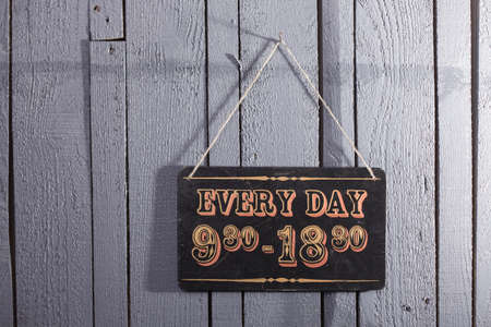 Closeup of one door plate or tablet of brown wood with time information for visiting hanging on rope indoor in studio on gery wooden backdrop, horizontal pictureの写真素材