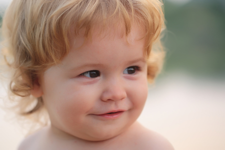 Closeup portrait of one cute funny playful happy smiling small boy with blonde curly hair and round cheeks looking away outdoor on natural background, horizontal pictureの写真素材