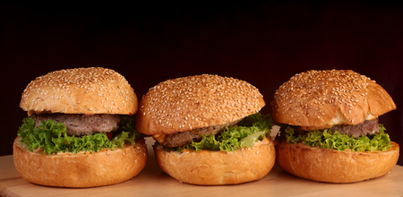 Big tasty appetizing fresh burgers of green lettuce red tomato cheese and bacon slice meat cutlet and white bread bun with sesame seeds closeup, horizontal pictureの写真素材