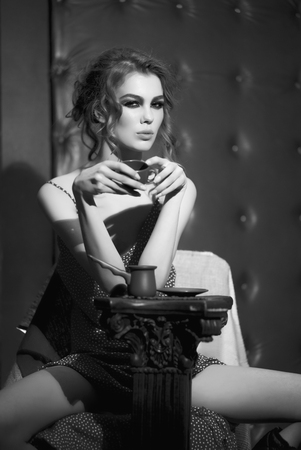 One beautiful sexy sensual young woman with bright makeup and curly hair in dress near column holding coffee cup sitting in studio on leather black background, vertical pictureの写真素材