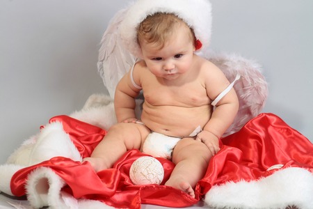 One small beautiful blonde boy child with curly hair in angel wings with feathers red hat and coat of santa playing with traditional decorating new year tree balls sitting in studio on grey backgroundの写真素材