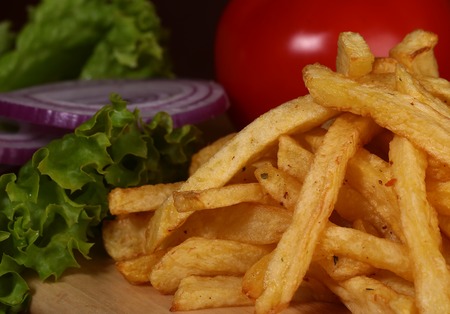 Closeup of fried fresh tasty potato chips sticks golden color lying in heap near red tomato violet onion and green lattuce on wooden background, horizontal pictureの写真素材