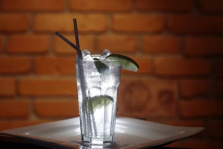 Long drink gin tonic cocktail with ice cubes and slices of lime in glass with two straws standing on...