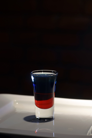 Closeup photo of one colorful twolevelled alcoholic shot cocktail red and blue colors in drinking glass standing on white plate on black background, vertical pictureの写真素材