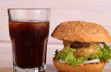 Big tasty appetizing fresh burger of green lettuce red tomato cheese and bacon slice meat cutlet and white bread bun with sesame seeds and glass of cola, horizontal pictureの写真素材