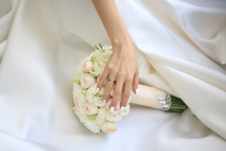 Beautiful fresh soft wedding decorative bouquet of white peony and rose flowers lying on dress of bride with female hand closeup, horizontal pictureの写真素材
