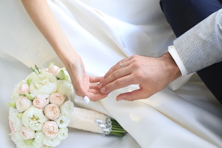 Beautiful fresh soft wedding decorative bouquet of white peony and rose flowers lying on dress of bride with female and male hands closeup, horizontal pictureの写真素材