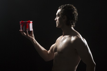 Side view of one handsome sexy strong muscular young man with bare chest and curly hair standing holding round red and white present box indoor in studio on black background, horizontal pictureの写真素材
