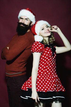New year funny couple of blond woman with curly hair and man with long beard in red santa claus hat celebrating christmas standing on studio purple background, verticalの写真素材