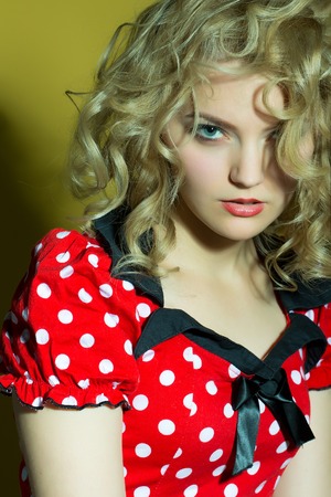 Portrait of one oung beautiful woman with blond curly hair in red white and black dress looking forward standing on studio yellow background, vertical pictureの写真素材