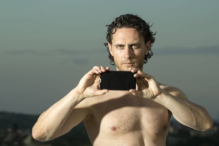 One young handsome serious unshaven muscular strong man with curly hair bare chest holding black mobile phone looking forward outdoor sunny day on blue sky natural background, horizontal pictureの写真素材