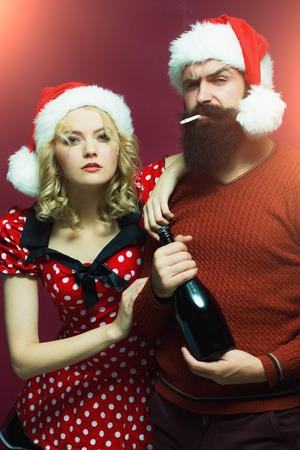 New year couple of blond woman with curly hair and man with long beard in red santa claus hat smoking cigarette holding wine bottle celebrating christmas standing on studio purple background, verticalの写真素材