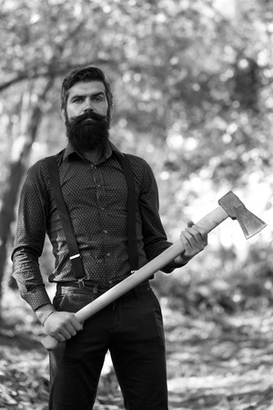 One serious man with long lush beard and moustache in shirt holding wooden axe standing sunny day outdoor on natural background black and white, vertical pictureの写真素材