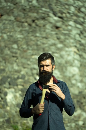 One handsome strong stylish male logger of young man with long lush black beard and moustache in shirt holding wooden axe standing near grey stone wall outdoor, vertical pictureの写真素材