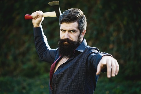 One handsome strong stylish male logger of young man with long lush black beard and moustache in shirt holding wooden axe standing near wall with climber plant outdoor, horizontal pictureの写真素材