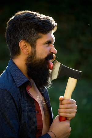 Side view of one handsome strong stylish male logger of young man with long lush black beard and moustache in shirt holding wooden axe standing near wall with climber plant outdoor, vertical pictureの写真素材