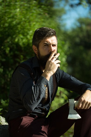 One strong stylish male logger of young man with long lush black beard and moustache in shirt holding wooden axe sitting in forest smoking cigarette outdoor on natural background, vertical pictureの写真素材