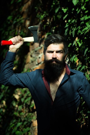 One handsome strong stylish male logger of young man with long lush black beard and moustache in shirt holding wooden axe standing near brick wall outdoor, vertical pictureの写真素材