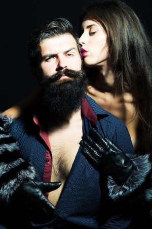 Beautiful stylish young sensual couple of woman in fur sleeves and gloves embraces man with long lush beard in shirt with bare torso sitting in studio on black background, vertical pictureの写真素材