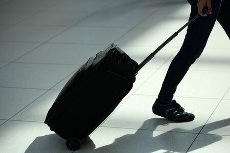 Closeup back view of female legs in jeans with black rolling suitcase wheeled travel bag trolley case moving over airport terminal floor tile background, horizontal pictureの写真素材