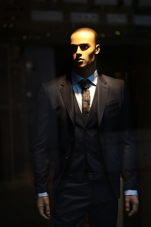 Closeup illuminated fashion mannequin in elegant black male suit made of unornamented cloth jacket pants vest tie of flowered fabric belt shirt in shop window over blur background, vertical pictureの写真素材