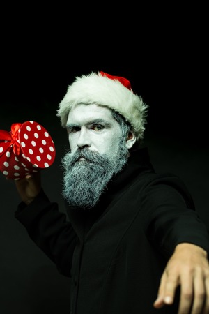 Closeup portrait of one senior frozen new year man with painted face in white and long beard in christmas santa red hat holding present box in studio on black background, vertical pictureの写真素材