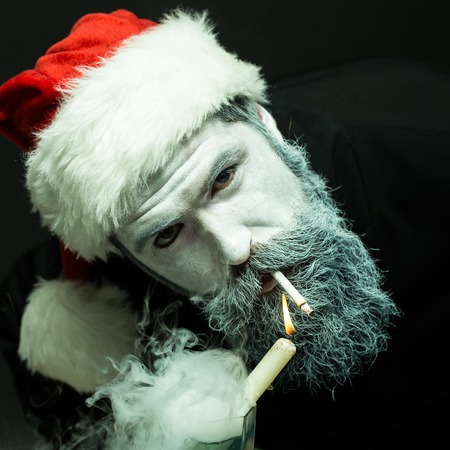 Closeup portrait of one senior frozen new year man with painted face in white and long beard in christmas santa red hat holding candle and smoking cigarette in studio on black background, verticalの写真素材
