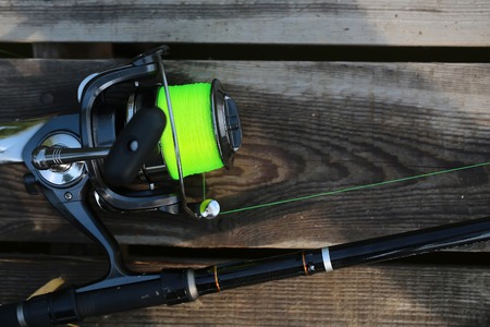 Closeup silhouette of fish-tackle fishing spinning rod and reel with  illuminative yellow colored angling line over natural wooden cover background, horizontal pictureの写真素材