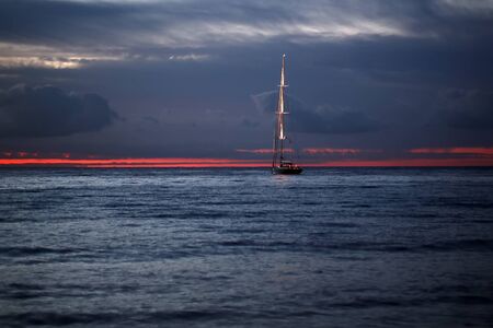 Beautiful calm evening seascape with yacht sailing in dark blue seaways under bare poles floating in the sea in rays of amazing red sunset on horizon against red and blue cloudy sky, horizontal photoの写真素材