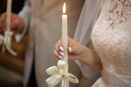 Beautiful bride in laced dress and groom holding burned candles decorated by white ribbon bow at wedding ceremony in church closeup indoor, horizontal pictureの写真素材