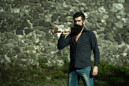 One handsome strong stylish male logger of young man with long lush black beard and moustache in shirt holding wooden axe standing near grey stone wall outdoor, horizontal pictureの写真素材