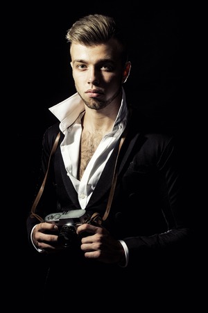 One young handsome sensual unshaven bearded man looking at camera with popped unbutton collar white shirt black jacket keeping camera stays forward in studio on black background, vertical pictureの写真素材