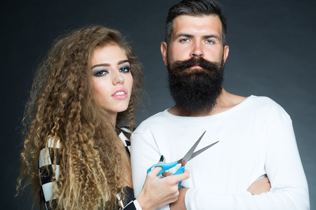 Couple of long-haired young beautiful woman holding scissors with handsome bearded gray-haired man with moustache looking forward on grey background, horizontal pictureの写真素材
