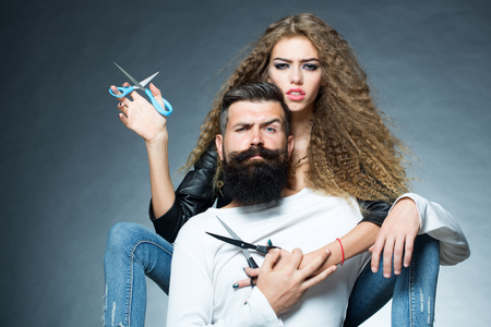 Couple of long-haired young beautiful woman holding two pairs of scissors sitting behind handsome bearded grey-haired man with moustache both looking forward on grey background, horizontal picture