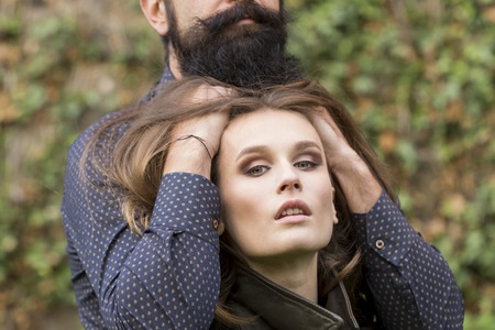 Young attractive woman with bright makeup and man with black beard touching her long lush hair standing outdoor on green natural background, horizontal pictureの写真素材