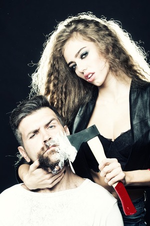 Portrait closeup couple of long-haired young sensual woman shaving handsome bearded grey-haired man with chopper and foam looking forward on grey background, vertical pictureの写真素材