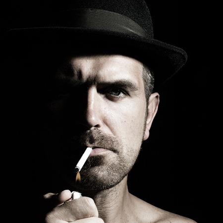 Portrait closeup young handsome sensual unshaven frown bearded man model with cigarette wearing felt hat studio play of light and shadow on black background, square pictureの写真素材