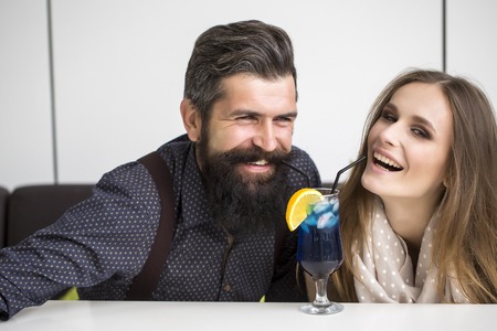 Closeup view on one attractive stylish emotional couple of young woman and senior man with long black beard drinking blue cocktail in glass with straw and orange slice intdoor, horizontal photoの写真素材