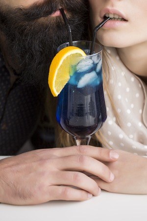 Closeup view on one attractive stylish emotional couple of young woman and senior man with long black beard drinking blue cocktail in glass with straw and orange slice sitting intdoor, vertical photoの写真素材