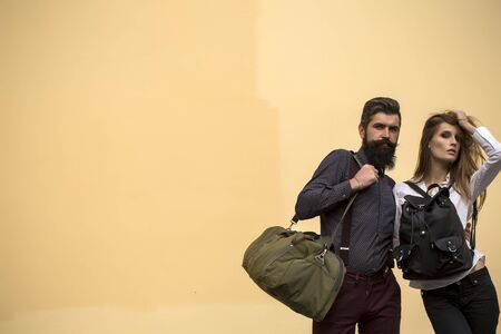 One beautiful stylish couple of young woman and senior man with long black beard standing embracing close to each other with travel bags outdoor in autumn street on light orange wall background, horizontal pictureの写真素材