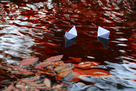 Closeup view of many beautiful colorful autumn tree leaves red yellow orange green colors floating on wavy water with reflection of nature with white paper ship on outdoor background, horizontal pictureの写真素材