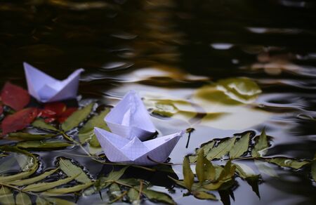 Closeup view of many beautiful colorful autumn tree leaves red yellow orange green colors floating on wavy water with reflection of nature with white paper ship on outdoor background, horizontal pictureの写真素材