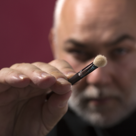 Closeup of one professional male visagiste with one natural makeup brush for eyeshadow powder and facial foundation doing makeup on purple background, square pictureの写真素材