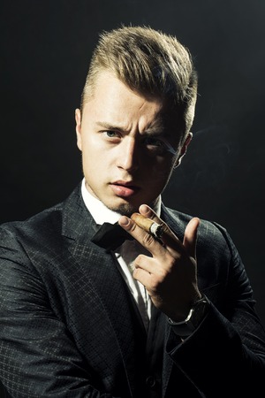 Portrait of presentable bold enchanting beautiful blonde boy in stylish formal suit and bow-tie holding cigar looking straight studio on black background closeup, vertical pictureの写真素材
